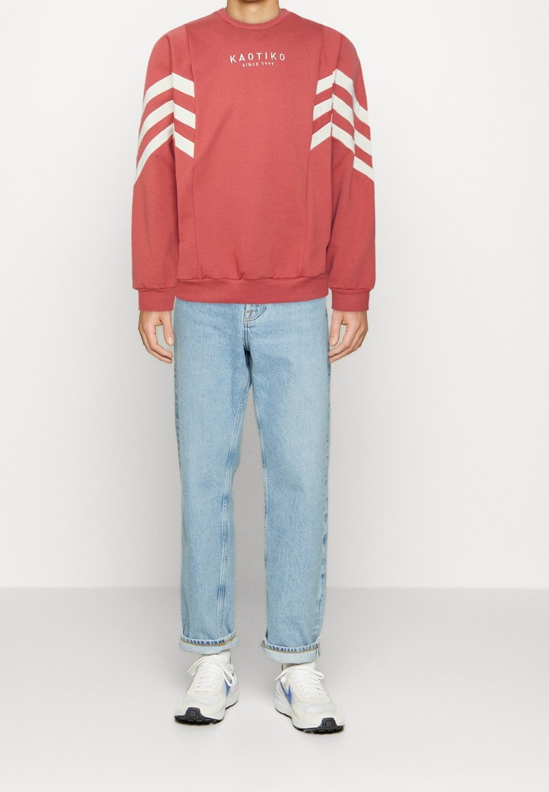 Personne portant un sweat-shirt rouge avec des rayures blanches sur les manches, un jean bleu et des baskets blanches, debout devant un fond uni.