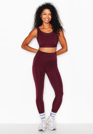 Donna con lunghi capelli ricci sorridente, indossa un crop top sportivo color marsala e leggings, scarpe da ginnastica bianche e calzini a righe, in piedi su sfondo bianco.