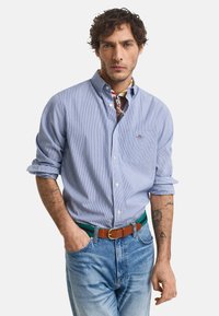 Vaaleansininen raidallinen nappipaita, jossa on tasku ja käännetyt hihat, yhdistetty haalistuneisiin sinisiin farkkuihin ja ruskeaan vyöhön, jossa on vihreitä yksityiskohtia.