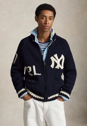 Młody mężczyzna z rękami w kieszeniach, ubrany w granatowy, dziergany kardigan na suwak z białym logo NY, warstwowo: denim i koszule w paski oraz białe spodnie.