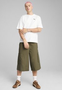 Vit kortärmad t-shirt med svart logotyp, olivgröna cargoshorts, vita strumpor och bruna sneakers med diskreta mönster och texturer.