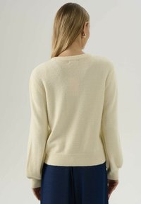 Donna con i lunghi capelli biondi, che indossa un maglione peloso color crema e pantaloni blu navy, vista di spalle su uno sfondo neutro.