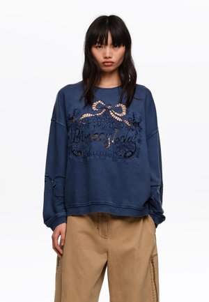 Joven con cabello largo y negro que lleva una sudadera azul marino con un lazo de encaje recortado y texto, combinada con pantalones sueltos de color marrón claro.