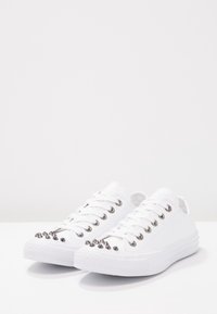 White canvas sneakers with a low-top design, featuring silver spikes on the toe, metal eyelets, flat laces, and a textured rubber sole.