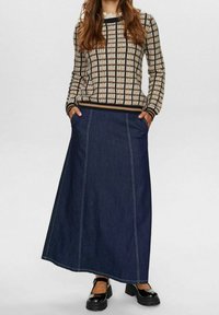 Jupe longue en denim avec poches latérales, complétée par un pull en maille à motifs beige et marron. Chaussures noires à talons épais viennent parfaire la tenue.