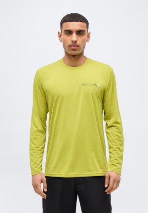 Mees, kes kannab laimirohelise pika varrukaga särki, millel on rinnal "NORRØNA" logo, seisab ühetaolise heleda tausta ees.