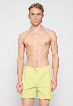 Joven hombre sin camiseta con cabello claro, vistiendo pantalones cortos de baño amarillos, de pie contra un fondo liso gris claro.