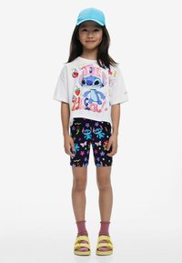 Witte cropped t-shirt met kleurrijke cartoonafbeelding, zwarte shorts met bloemen- en cartoonpatronen, roze sokken en gele sandalen.