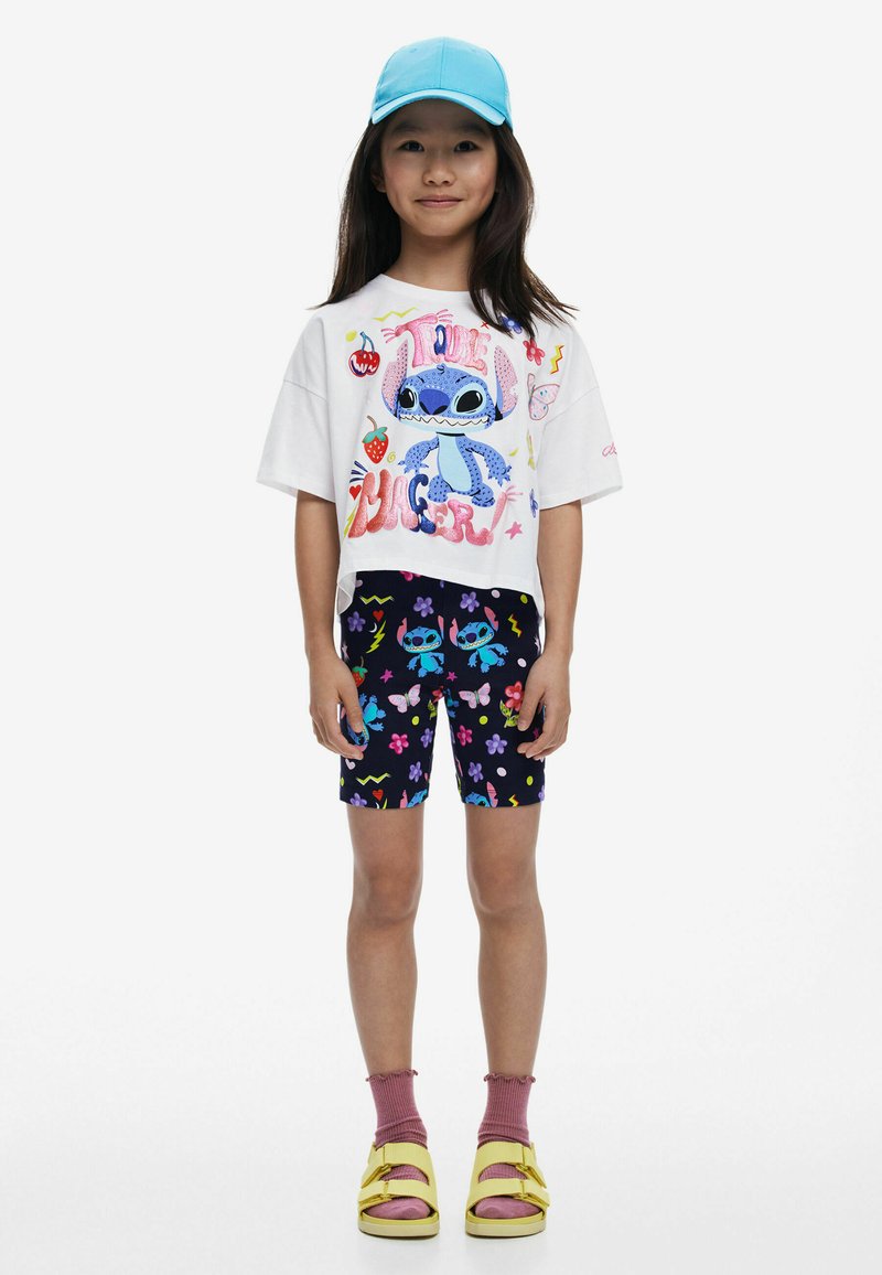 Witte cropped t-shirt met kleurrijke cartoonafbeelding, zwarte shorts met bloemen- en cartoonpatronen, roze sokken en gele sandalen.