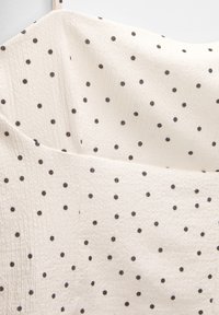 Hvid struktureret stof med små sorte prikker. Designet inkluderer en enkel, glat halsudskæring og subtile lag i stoffet.