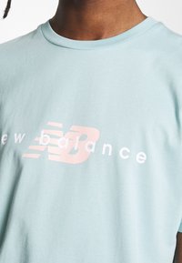 Světle modré bavlněné tričko s kulatým výstřihem; na hrudi má bílé a růžové logo "New Balance" v stylizovaném písmu.