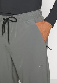 Šedé športové nohavice s elastickým pásom, nastaviteľnou šnúrkou, bočnými vreckami a nenápadným strieborným logom Nike. Hladká štruktúra.