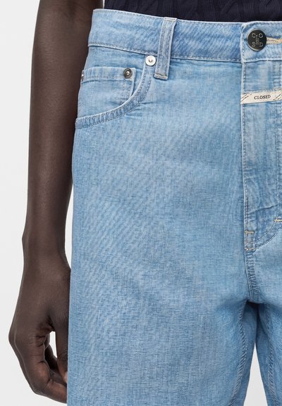 Φωτεινά μπλε σορτς τζιν με ψηλή μέση, μπροστινές τσέπες και κουμπί και ετικέτα με το λογότυπο "Closed", φορεμένα από άτομο με σκούρο δέρμα.