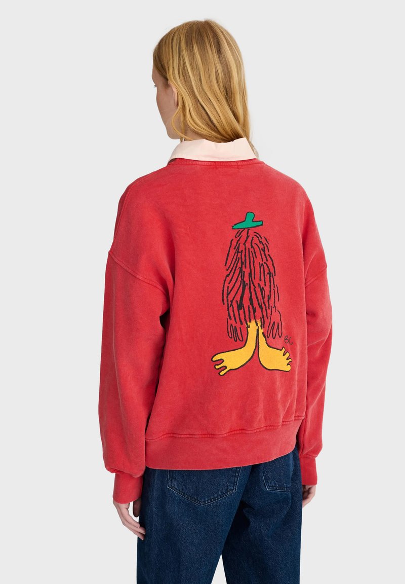 Personne blonde portant un sweat-shirt rouge avec un dessin animé représentant une silhouette poilue aux pieds jaunes et au chapeau vert dans le dos, associé à un jean bleu.