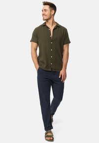 Short-sleeve, olive green shirt with a textured finish and button front, paired with navy pants and green sandals. Model standing and smiling.