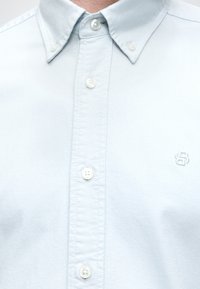 Chemise bleu clair à boutons blancs avec un petit logo brodé sur la poitrine gauche.