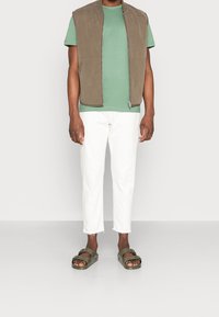 Kaki ärmlös väst över en grön t-shirt, kombinerat med vita capribyxor och olivgröna sandaler. Mjuka texturer och avslappnad passform.