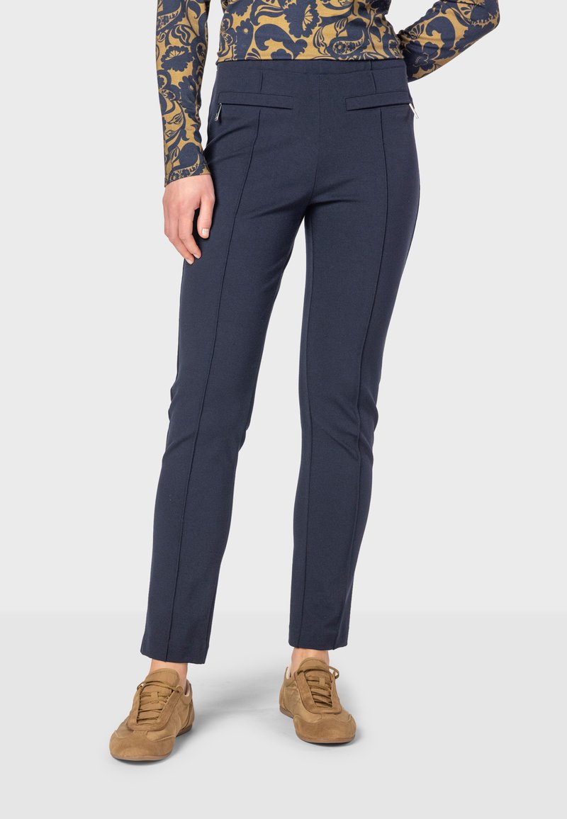 Navy blue trousers with a slim fit, featuring front pockets with zippers, and vertical seam detailing; paired with light brown sneakers.