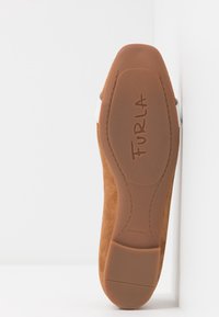 Semelle de chaussure en daim beige avec une base en caoutchouc texturé, présentant un logo "FURLA" bien en vue, ainsi qu'un détail de sangle blanche.