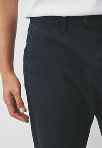 Dark navy chinos made of cotton blend, with a smooth texture, slim fit, side pockets, and a button closure. Visible hand near waistband.