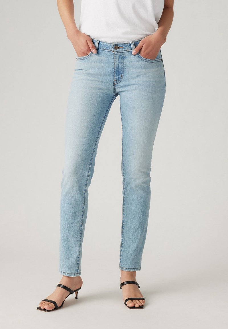 Femme portant un jean slim bleu clair et des sandales à talons noires à bouts ouverts, debout les mains dans les poches devant un fond uni.