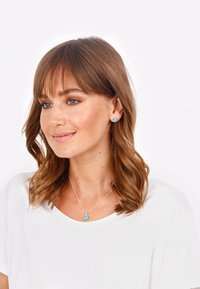 Collana d'argento con pendente in turchese; orecchini a bottone coordinati in turchese; modella che indossa una t-shirt bianca; capelli castani morbidi e mossi.