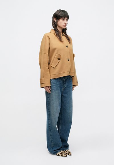Veste marron à double boutonnage avec grandes poches avant, associée à un jean bleu à jambes larges et des chaussures imprimé léopard. Fond blanc uni.