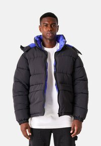 Zwarte gewatteerde jas met blauwe voering, voorzien van een ritssluiting en elastische manchetten. De jas heeft een capuchon en zichtbare stikseldetails.