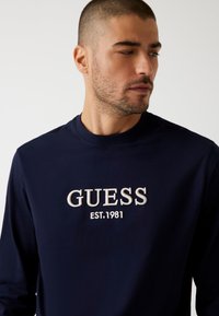 Felpa blu navy con scollo rotondo, caratterizzata da un logo "GUESS" ricamato in bianco e "EST. 1981" sottostante. Texture morbida, design casual.