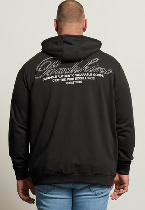 Homme portant un sweat à capuche noir avec le texte "Badrhino" et un slogan au dos, associé à un jean bleu clair, debout dos à la caméra.