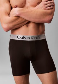 Brązowe bokserki z srebnym paskiem z logo Calvin Klein. Materiał wygląda na gładki, ma dopasowany krój i długość do połowy uda.