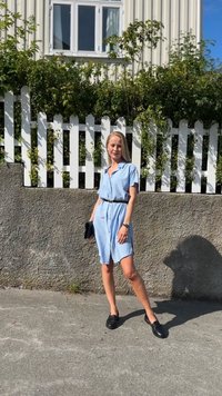 Hellblaues Hemdkleid mit kurzen Ärmeln, einem taillierten Bund und Taschen; kombiniert mit schwarzen Slippern. Der Hintergrund zeigt Grünanlagen und einen weißen Zaun.