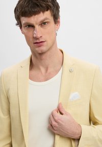 Helles gelbes Blazer mit strukturierter Oberfläche, einzelner Blumenanstecker und weißem Taschentuch; über einem weißen gerippten Tanktop getragen.