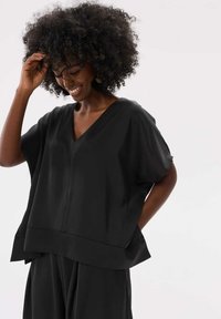 Zwarte, loszittende top met een V-hals, brede mouwen en een zoom die op de heupen valt. Soepele textuur met minimale stikseldetails.