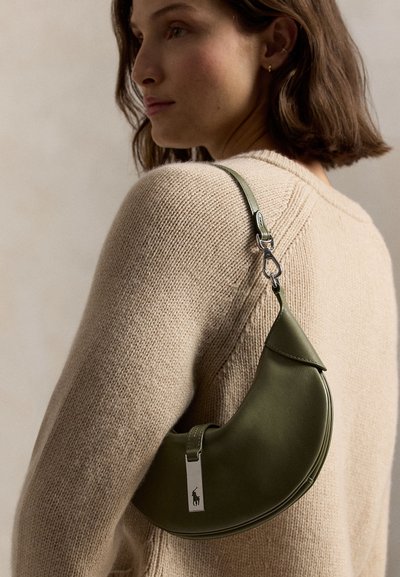 Mujer con cabello castaño hasta los hombros, vestida con un suéter de punto beige, llevando un bolso de cuero verde en forma de media luna con cierre plateado.