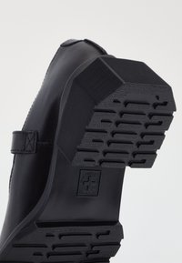 Black leather shoe with a textured rubber sole, featuring a hexagonal design and a visible logo. The heel is angular with horizontal grooves.