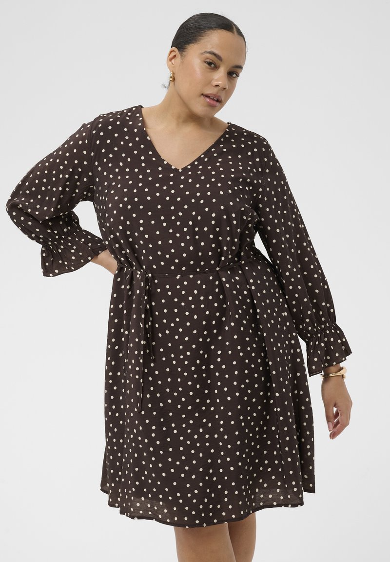 Femme portant une robe noire à manches longues avec des pois blancs, mains sur les hanches, debout devant un fond clair uni.