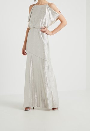 Femme portant une robe de soirée argentée, longue jusqu'au sol, aux épaules dénudées, fines bretelles et sandales à talons hauts, debout avec une main sur la hanche.
