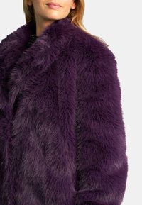 Abrigo de piel sintética morado con una textura rica y suave, que cuenta con un cuello y mangas amplias. La tela presenta una apariencia densa y esponjosa.