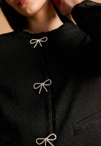 Čierna textúrovaná bunda s tromi uzávermi v tvare mašlí zdobenými kryštálmi, s vreckom viditeľným na ľavej strane.