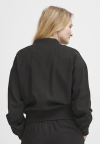 Čierna bomber bunda s priliehavým pásom a mäkkou tkaninou. Má vzpriamený golier a voľné rukávy pre pohodlie.