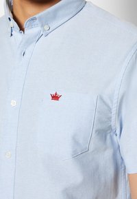 Camicia azzurra chiaro con bottoni, realizzata in tessuto morbido, con una tasca sul petto decorata da un ricamo di una corona rossa. Maniche corte, design del colletto classico.