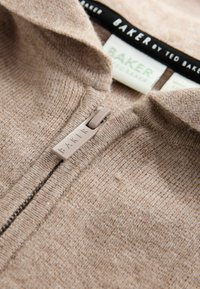 Beżowy sweter zapinany na zamek, wykonany z teksturowanej tkaniny, z czarno-białą metką kołnierza i jasnobeżowym uchwytem zamka z wytłoczonym napisem "BAKER".