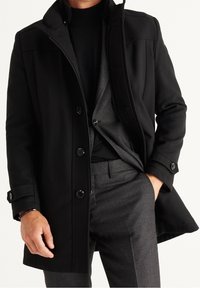 Cappotto nero con un design a colletto, caratterizzato da grandi bottoni e una texture liscia. Abbinato a pantaloni da completo grigi con texture.