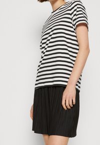 Junge Person trägt ein schwarz-weiß gestreiftes kurzärmeliges T-Shirt und einen schwarzen, plissierten Rock, steht vor einem schlichten Hintergrund.