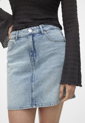 Femme portant une mini-jupe en denim bleu clair avec des poches devant et un haut noir texturé à manches longues avec des poignets évasés.