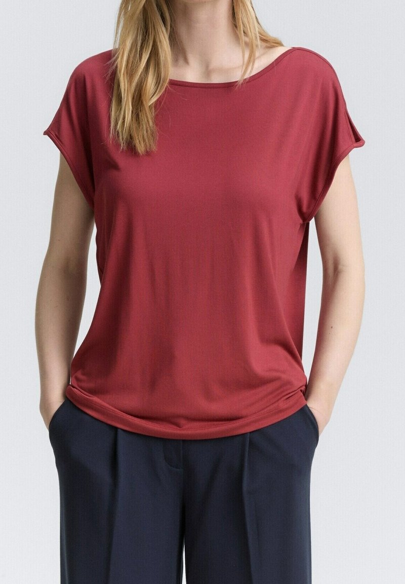 Haut bordeaux à manches courtes, coupe ample et encolure bateau, en tissu doux et lisse. Porté avec un pantalon bleu marine, mains dans les poches.