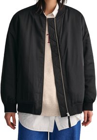 Blouson aviateur noir avec intérieur matelassé, poignets et ourlet côtelés, fermeture à glissière intégrale à l'avant et coupe décontractée. Porté sur un sweat léger et une chemise blanche.