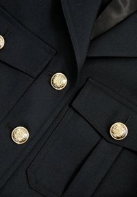 Čierna tailored bunda s zlatými gombíkmi v tvare leva, ostrými klopami a textúrovanou látkou, obsahujúca dva predné vrecká s klopou.