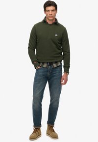 Man staat in een olijfgroene sweatshirt over een geruit shirt, blauwe jeans, tan veterschoenen en een polshorloge tegen een witte achtergrond.